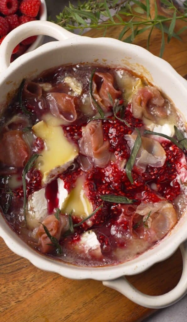 Baked brie with raspberry preserve and prosciutto freshly out of the oven, bubbling in a white ceramic dish