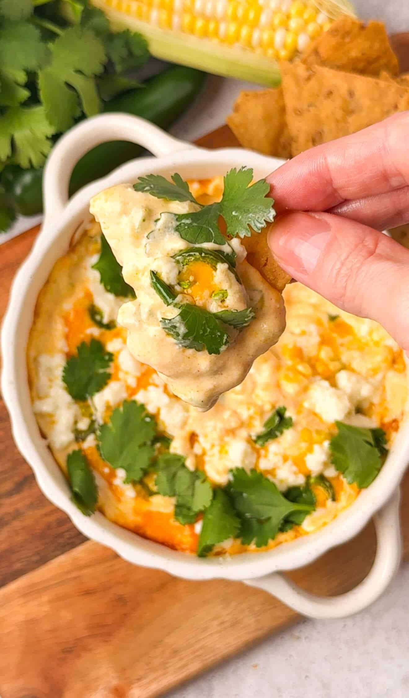Baked corn and jalapeño dip in a white ceramic dish with a tortilla chip being pulled through the creamy cheesy filling, topped with crumbled cotija and fresh cilantro