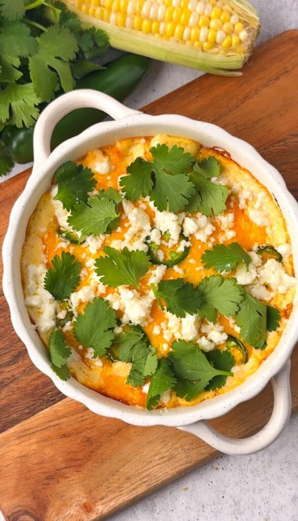 Overhead view of baked corn jalapeño dip in a white ceramic baking dish topped with melted cheddar, crumbled cotija, jalapeño slices, and fresh cilantro leaves on a wood board