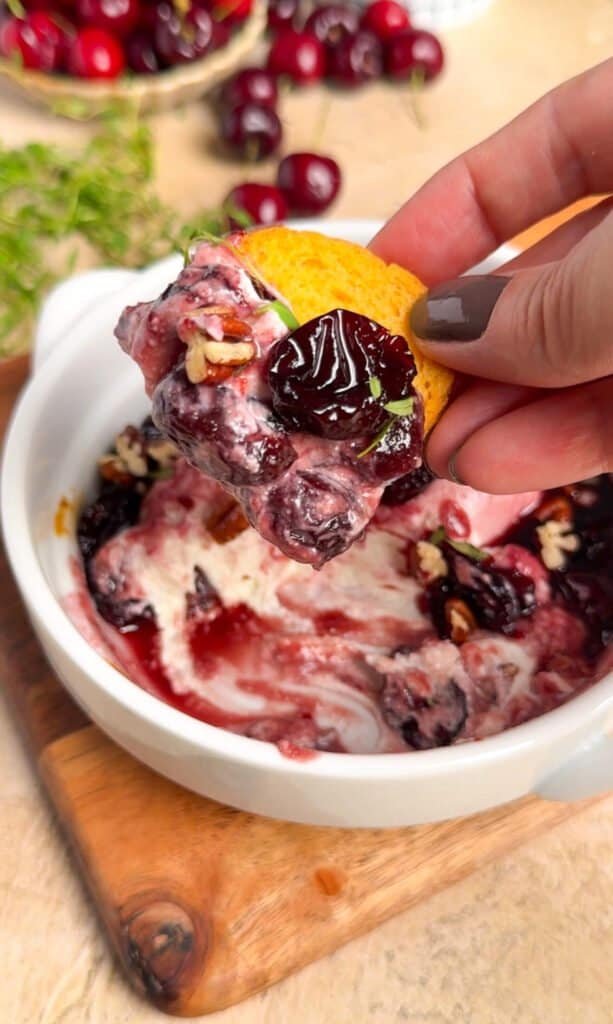 Hand scooping baked goat cheese and dark cherry jam onto a crostini from a white baking dish, toasted pecans and fresh cherries visible in background