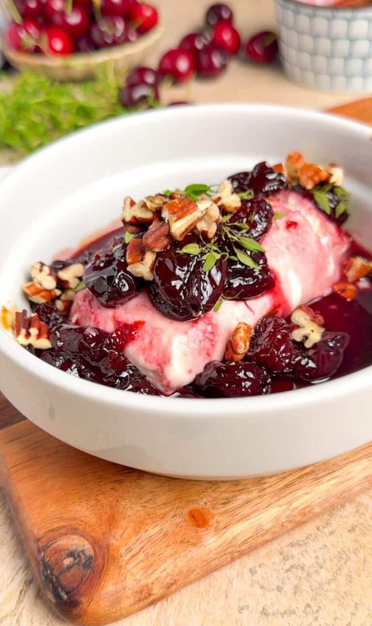 Baked goat cheese with dark cherry jam, toasted pecans, and fresh thyme in a white ceramic baking dish on a rustic wood board, fresh cherries in background
