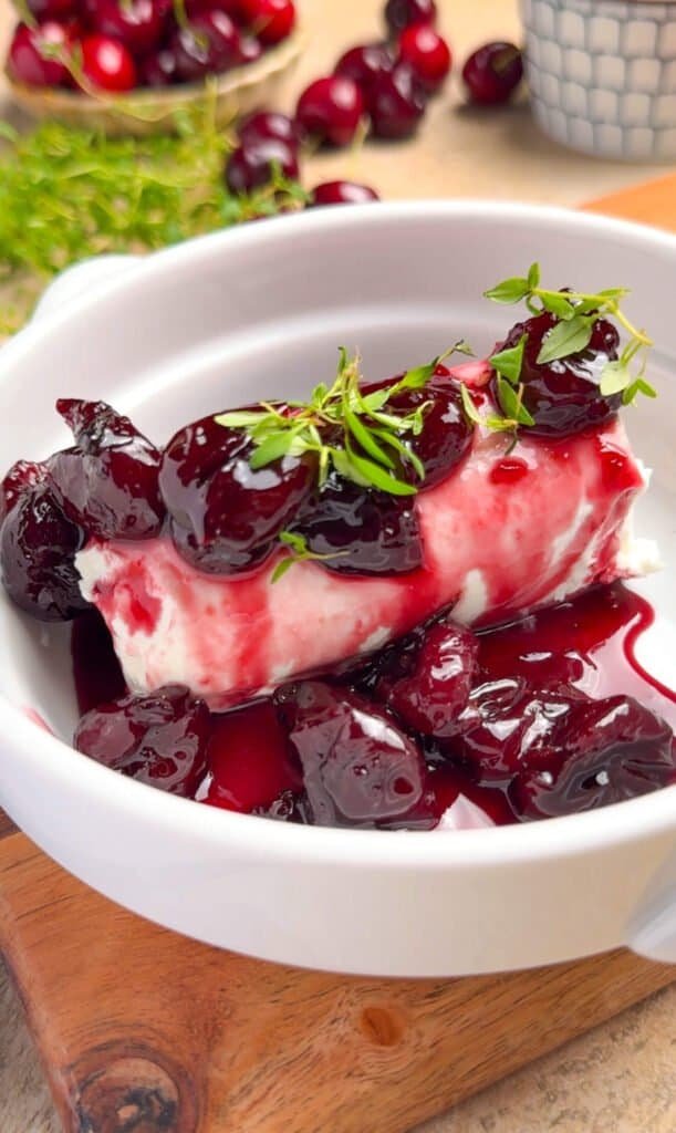 Baked goat cheese log nestled in bubbling dark cherry and red wine jam in a white ceramic baking dish, garnished with fresh thyme sprigs, on a wood serving board