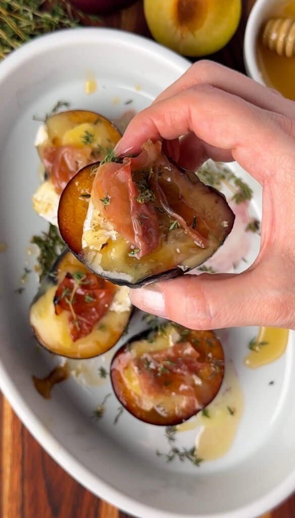 Hand picking up a baked plum half topped with Camembert, prosciutto, and thyme from a white baking dish