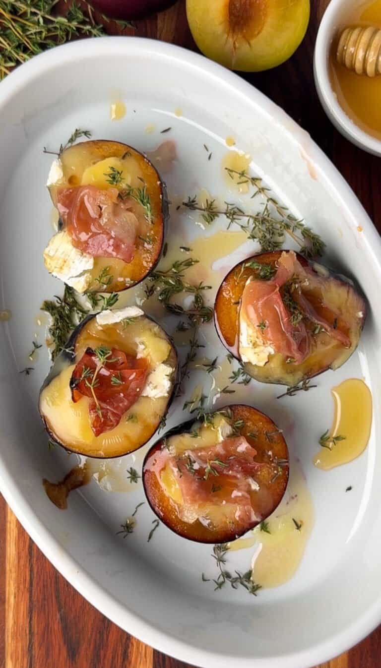 Four baked plum halves with Camembert, prosciutto, and fresh thyme in a white baking dish, drizzled with honey