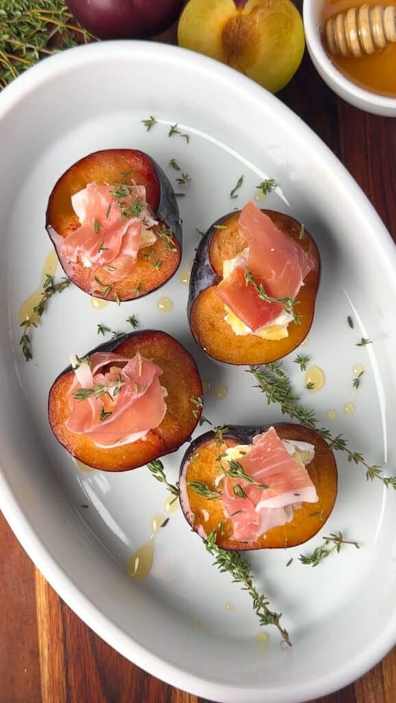 Overhead view of baked plums with melted Camembert, prosciutto, and thyme in a white oval dish, with fresh plums and a honey dipper in the background