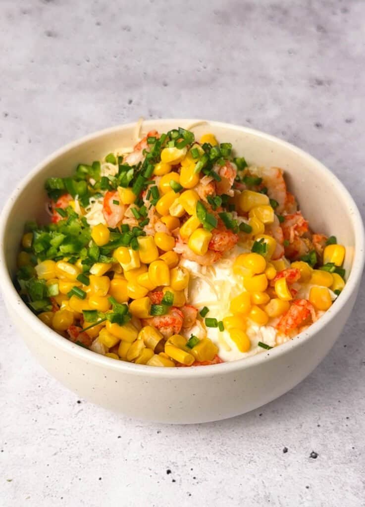 Bowl of crawfish cheesecake filling ingredients before mixing — crawfish tails, roasted corn, jalapeño, chives, smoked Gouda, and cream cheese in a white ceramic bowl