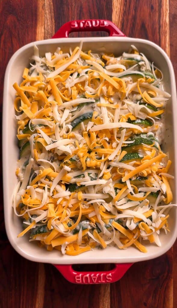 Freshly grated Swiss and mild cheddar cheese scattered over layered zucchini slices in a red Staub casserole dish before baking