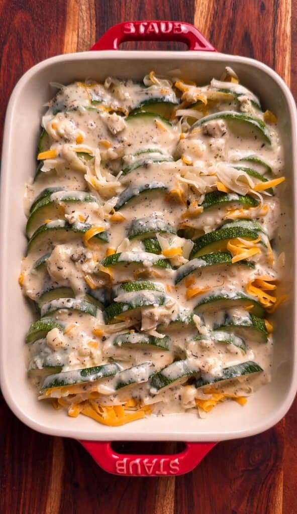 Brie cream sauce poured over overlapping zucchini slices and shredded cheddar and Swiss cheese in a Staub baking dish