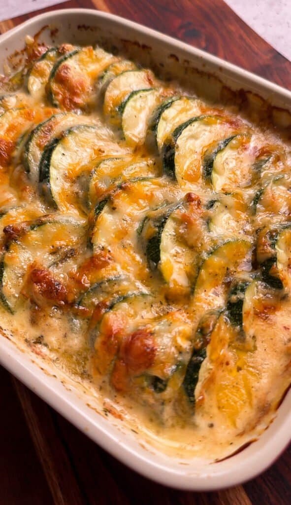 Cheesy zucchini casserole with a golden broiled cheese crust in a white ceramic baking dish on a wood cutting board