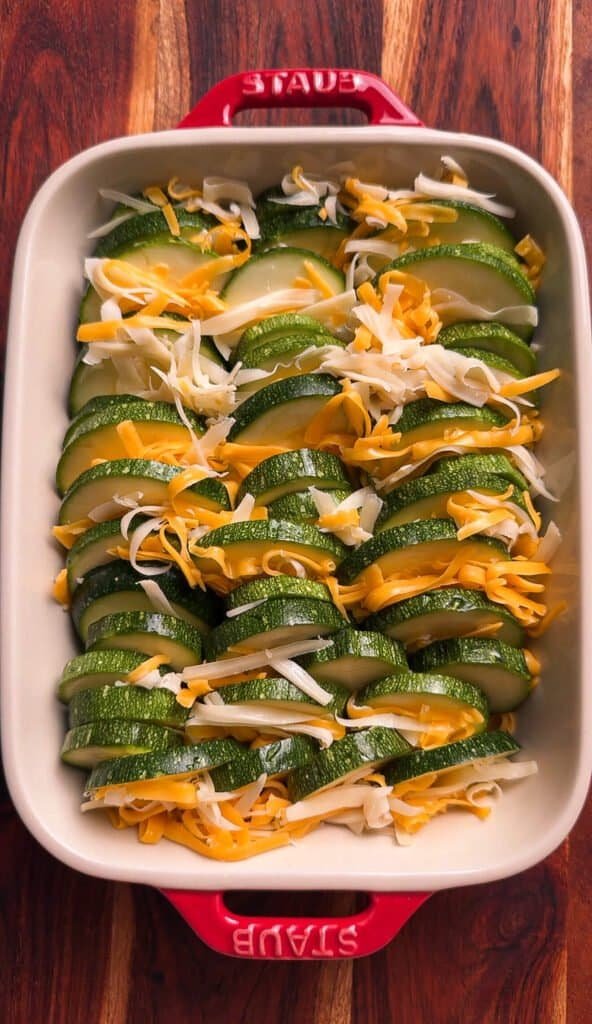 Raw zucchini rounds layered in overlapping rows with freshly grated Swiss and cheddar cheese in a red Staub casserole dish