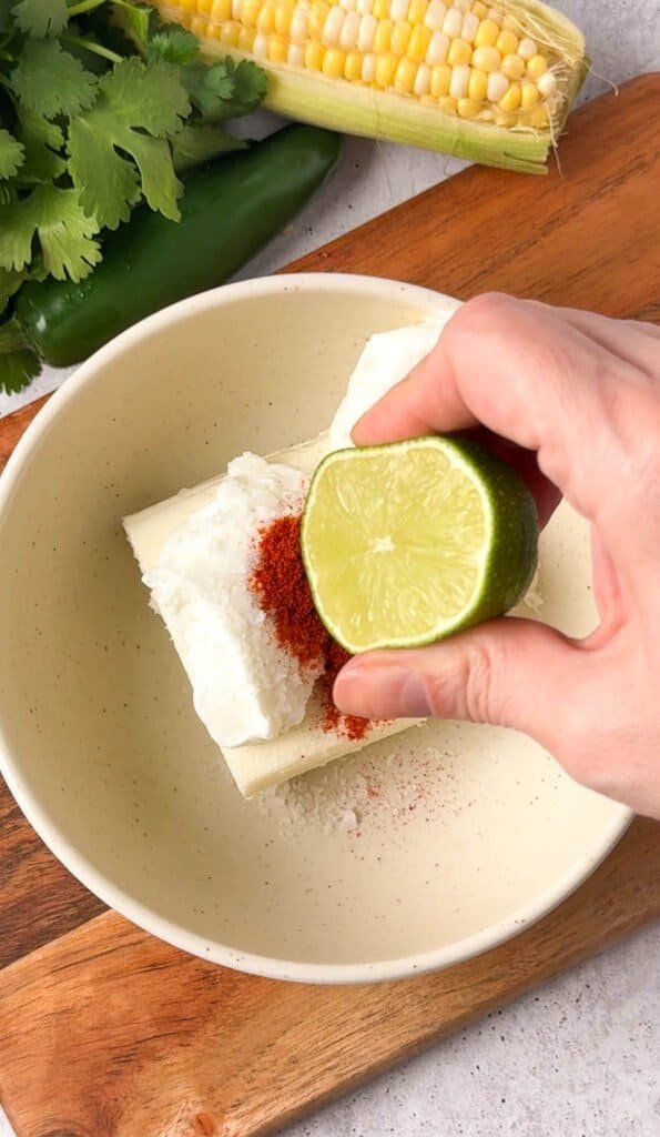 Hand squeezing fresh lime juice over softened cream cheese with smoked paprika and salt in a bowl for baked corn dip base