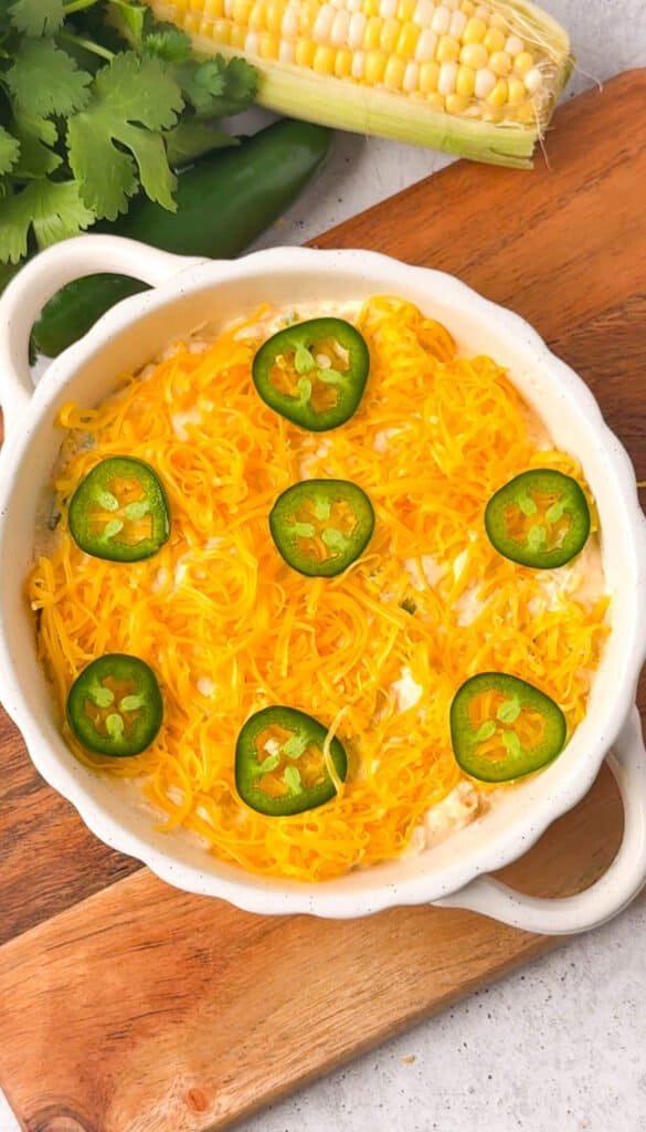 Unbaked corn jalapeño dip topped with shredded cheddar and fresh jalapeño slices in a white ceramic dish, ready to go into the oven