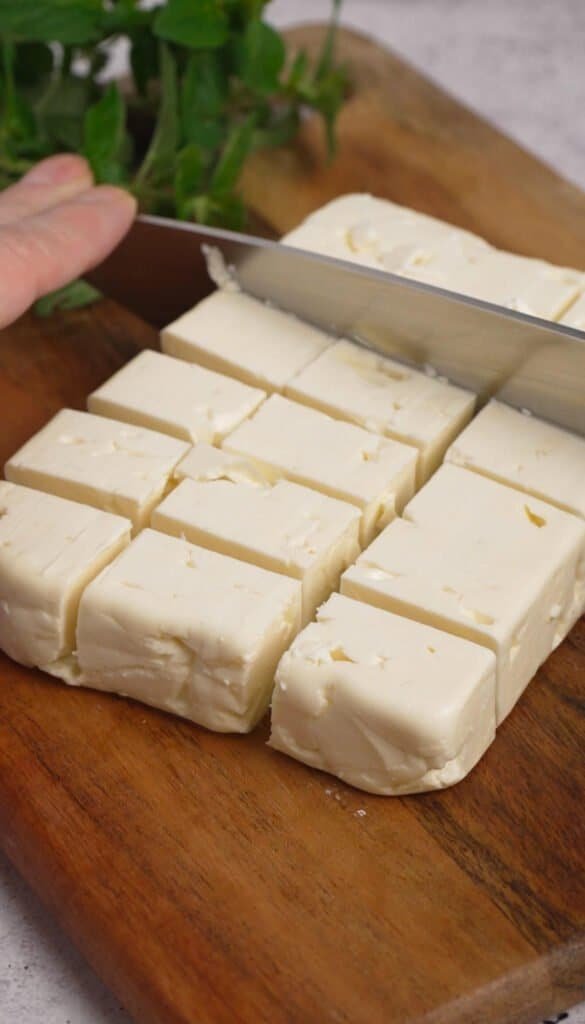 Cutting a block of feta cheese into cubes on a wooden cutting board with fresh herbs in the background