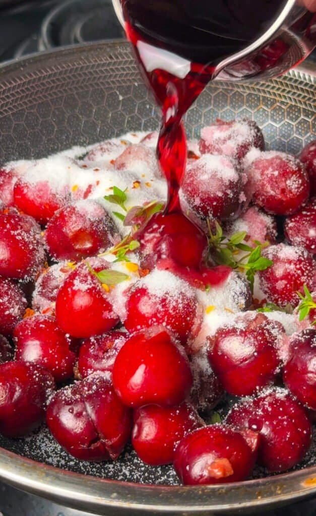 Dark sweet cherries dusted with sugar and fresh thyme sprigs in a skillet as red wine is poured over them to make small batch dark cherry jam