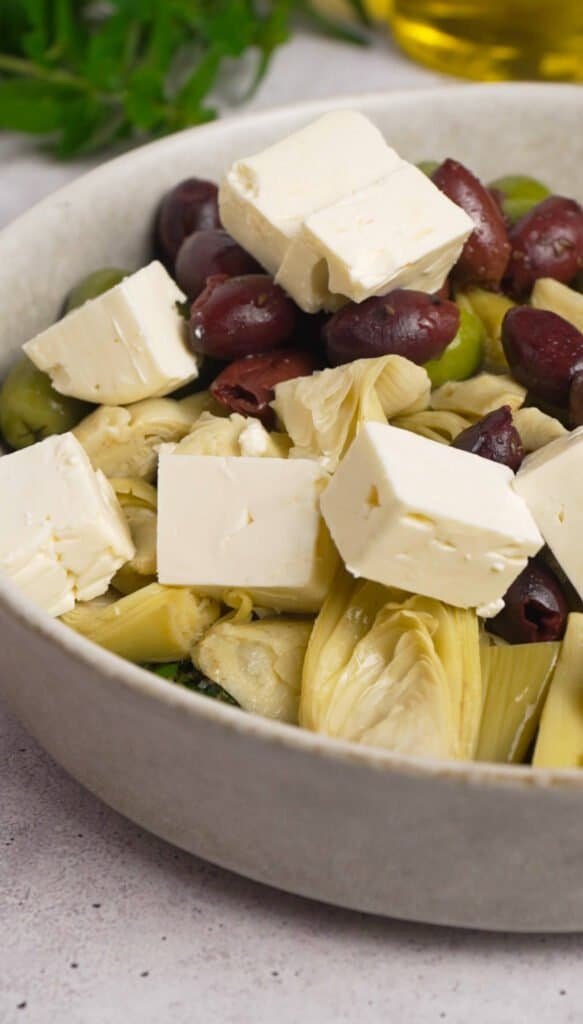 Feta cubes, quartered artichoke hearts, Kalamata and Castelvetrano olives added to herb marinade in a white bowl before tossing