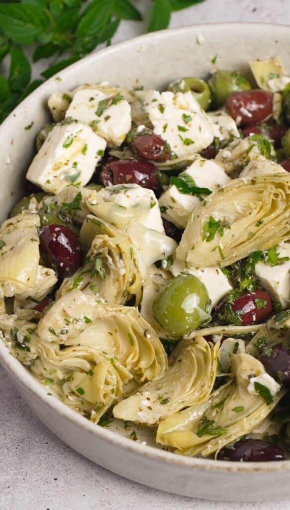 Marinated feta with artichoke hearts and mixed olives tossed in herb marinade, close-up in a white ceramic bowl