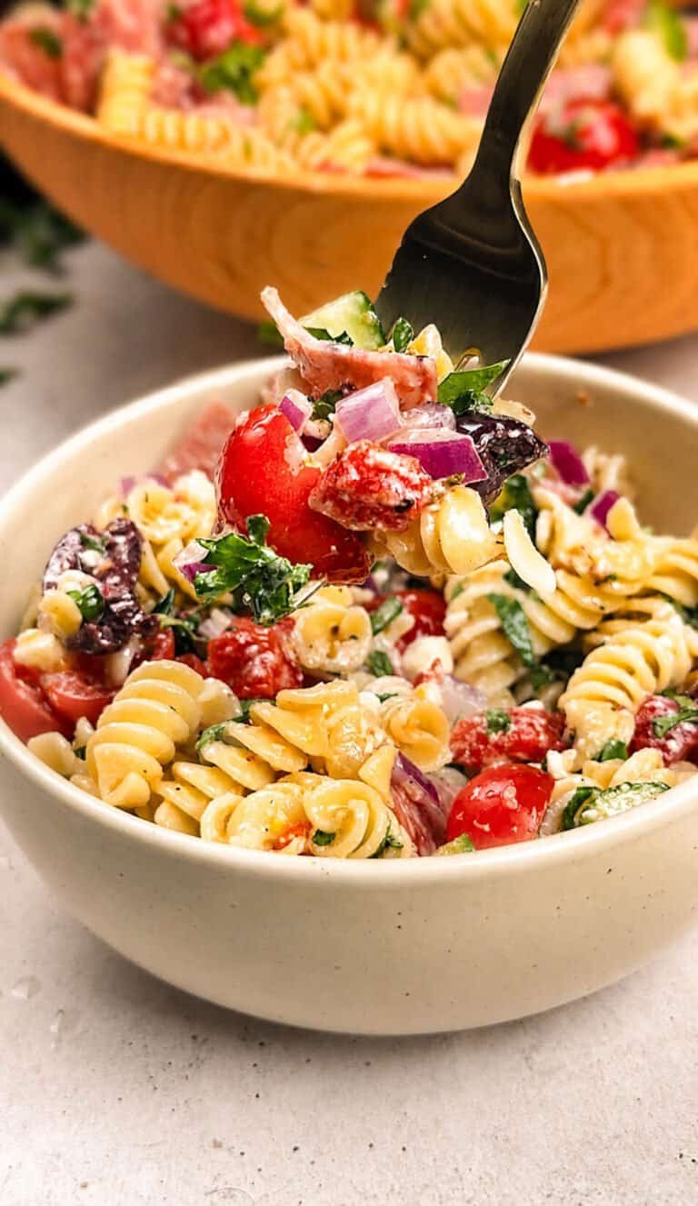 Mediterranean pasta salad in a white ceramic bowl with a fork lifting rotini, cherry tomatoes, red onion, and salami, with a wooden serving bowl in the background