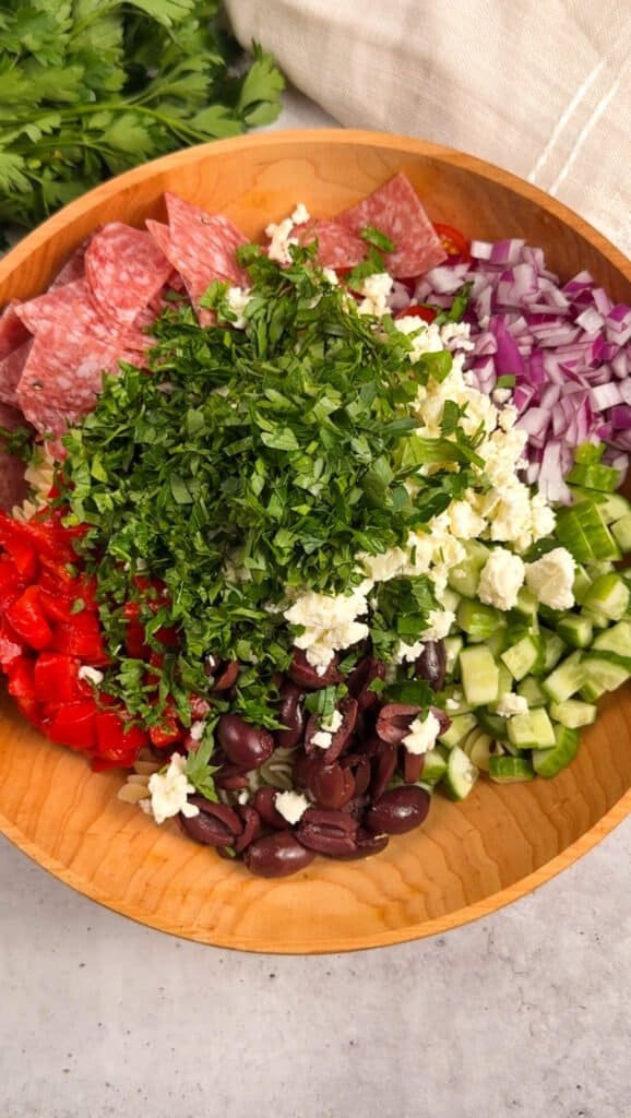 Mediterranean pasta salad ingredients arranged in a wooden bowl before tossing, including salami, crumbled feta, Kalamata olives, roasted red peppers, cucumber, red onion, and fresh herbs