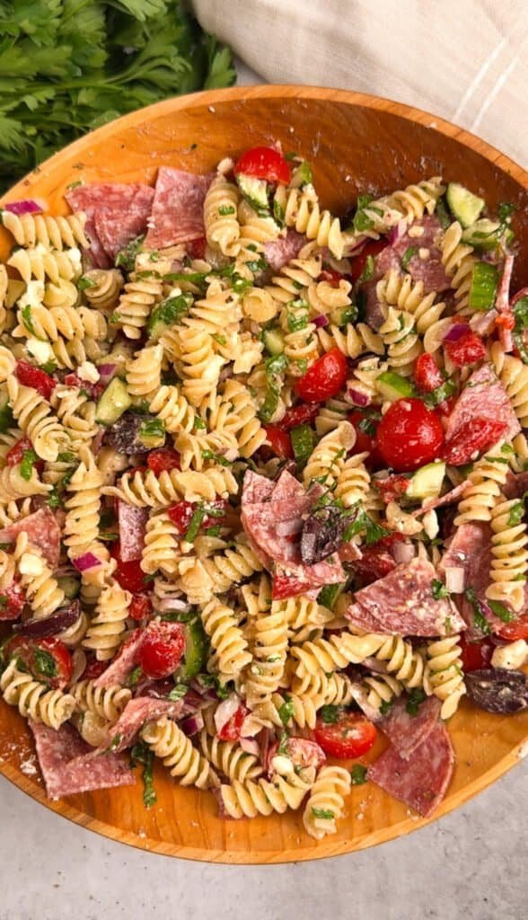 Overhead view of Mediterranean pasta salad in a large wooden bowl with rotini, salami, cherry tomatoes, Kalamata olives, red onion, cucumber, feta, and chopped parsley