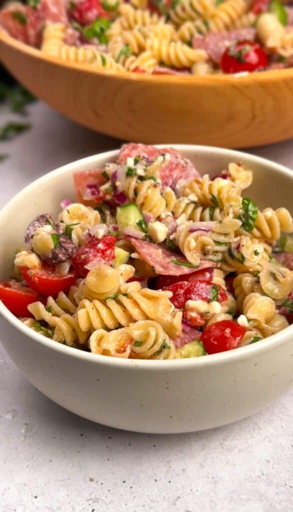 Mediterranean pasta salad served in a white speckled bowl with rotini, salami, cherry tomatoes, cucumber, Kalamata olives, feta, and fresh herbs