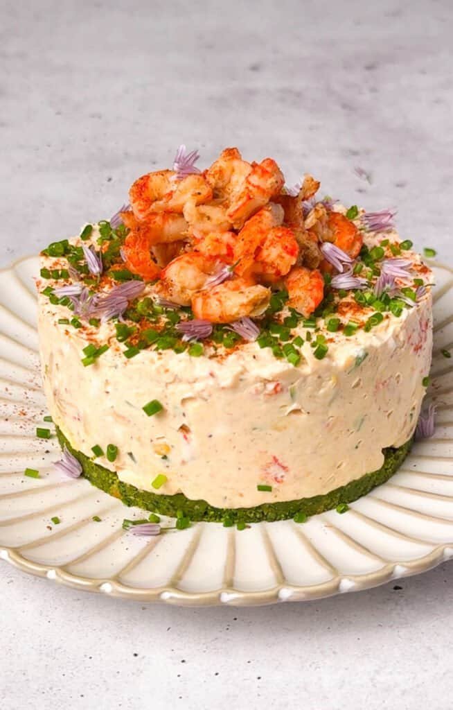 No-bake Cajun crawfish savory cheesecake on a white ridged ceramic plate, with a vivid green spinach cracker crust, textured cream cheese filling, pile of Cajun crawfish tails, chive blossoms, and smoked paprika dusting