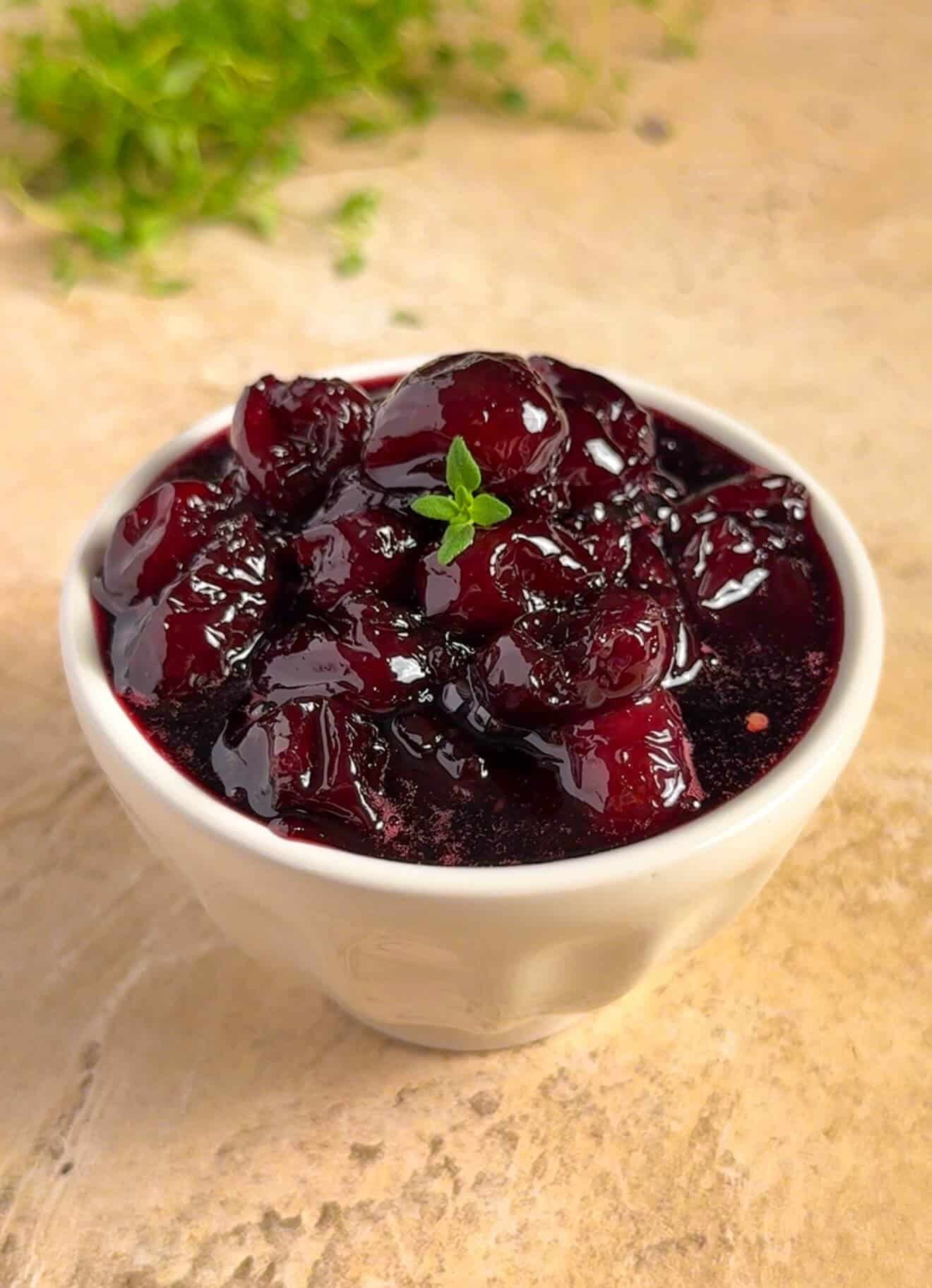 Small batch dark cherry and red wine jam with thyme served in a white ceramic bowl on a stone surface, garnished with a fresh thyme sprig