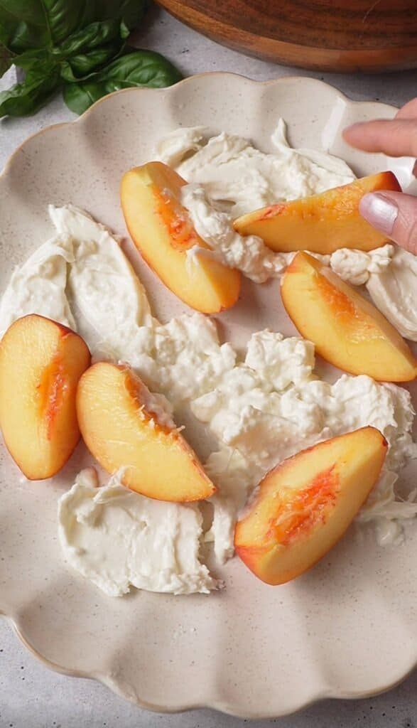 Torn burrata cheese and peach wedges arranged on a plate while assembling peach burrata salad