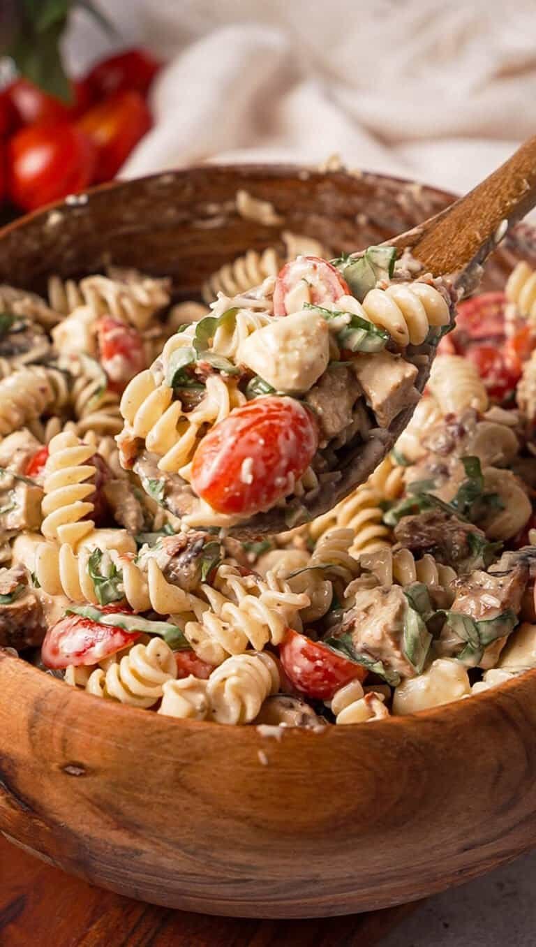 A wooden spoon lifting a scoop of creamy balsamic chicken pasta salad with basil, tomatoes, and mozzarella