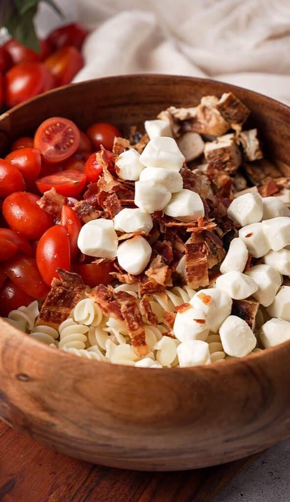 Cherry tomatoes, crispy bacon, mozzarella pearls, and chopped chicken arranged in a wooden bowl before mixing creamy balsamic chicken pasta salad