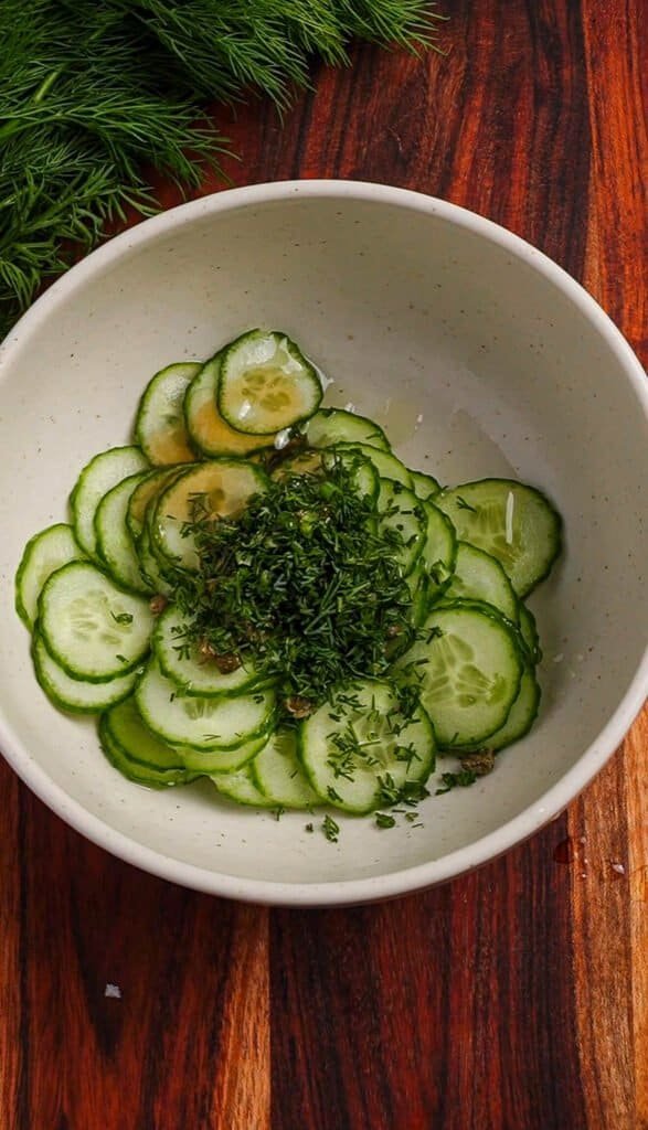 Thinly sliced Persian cucumber coins with fresh dill and herbs in a white bowl before tossing