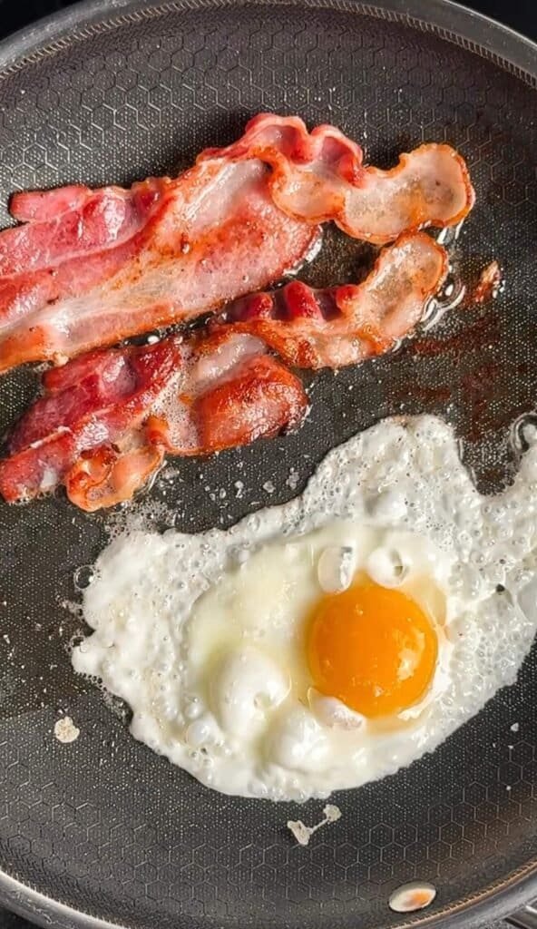 Bacon and a sunny side up egg frying together in a skillet for loaded avocado toast