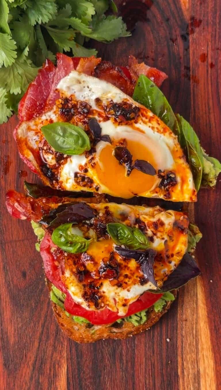 Loaded avocado toast sliced in half showing a runny egg yolk, crispy bacon, and chili crunch drizzle