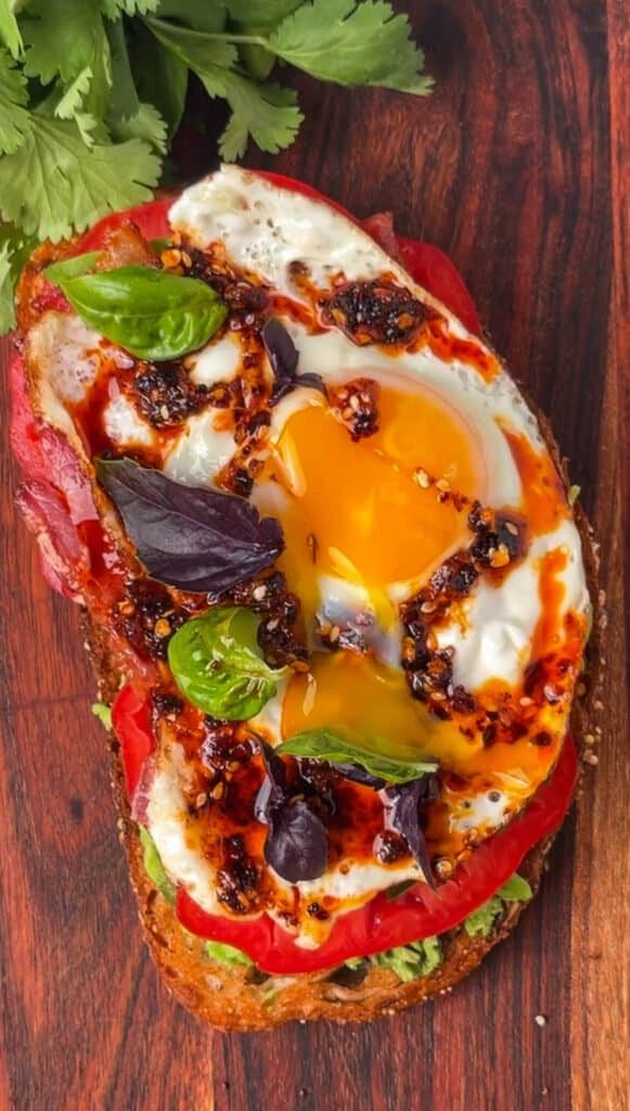 Close-up of loaded avocado toast with a broken egg yolk, purple basil, and chili oil on sourdough