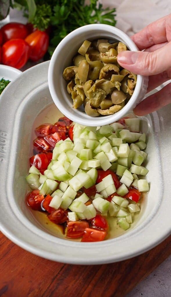 Chopped green olives being added to diced tomatoes and cucumber for Mediterranean tomato dip