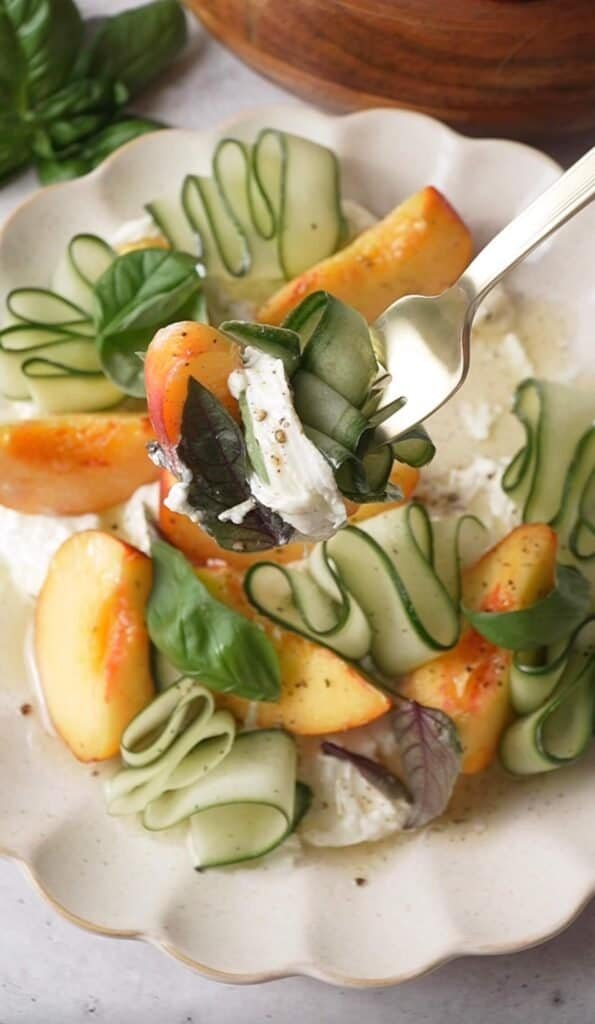 Forkful of peach burrata salad with cucumber ribbon, basil, and creamy burrata lifted from the plate
