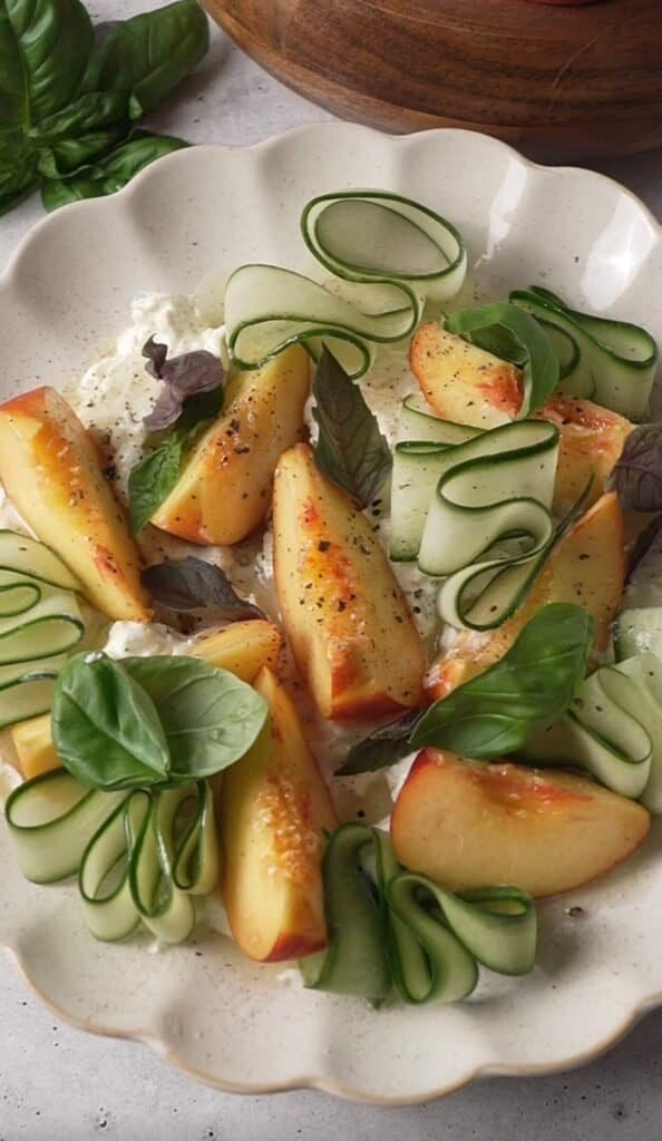 Overhead view of peach burrata salad with cucumber ribbons and fresh basil on a scalloped plate