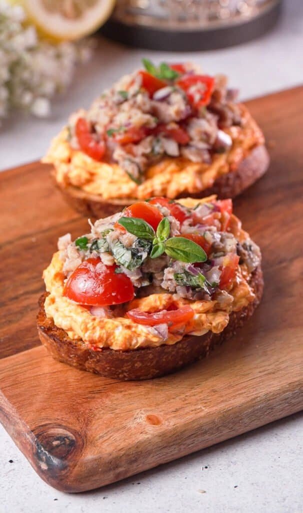 Two sardine toasts with harissa cream cheese and fresh tomatoes on a wood board