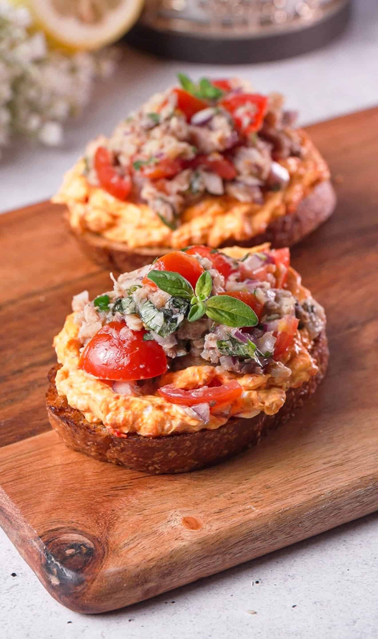 Two sardine toasts with harissa cream cheese and fresh tomatoes on a wood board