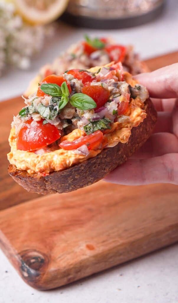 Hand holding a slice of sardine toast with harissa cream cheese and basil
