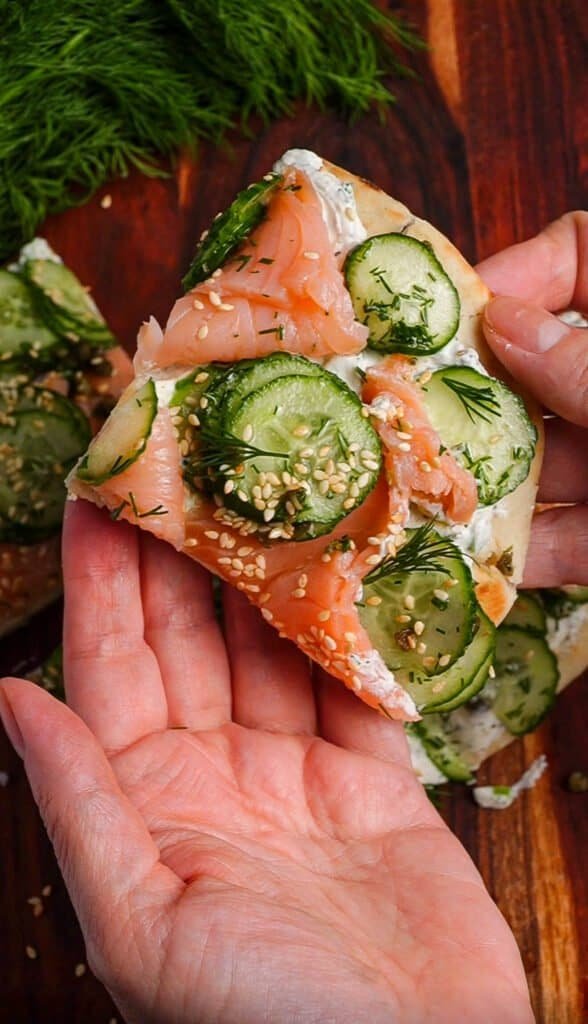 Hands holding a slice of smoked salmon flatbread with whipped herb labneh, cucumber caper salad, and sesame seeds