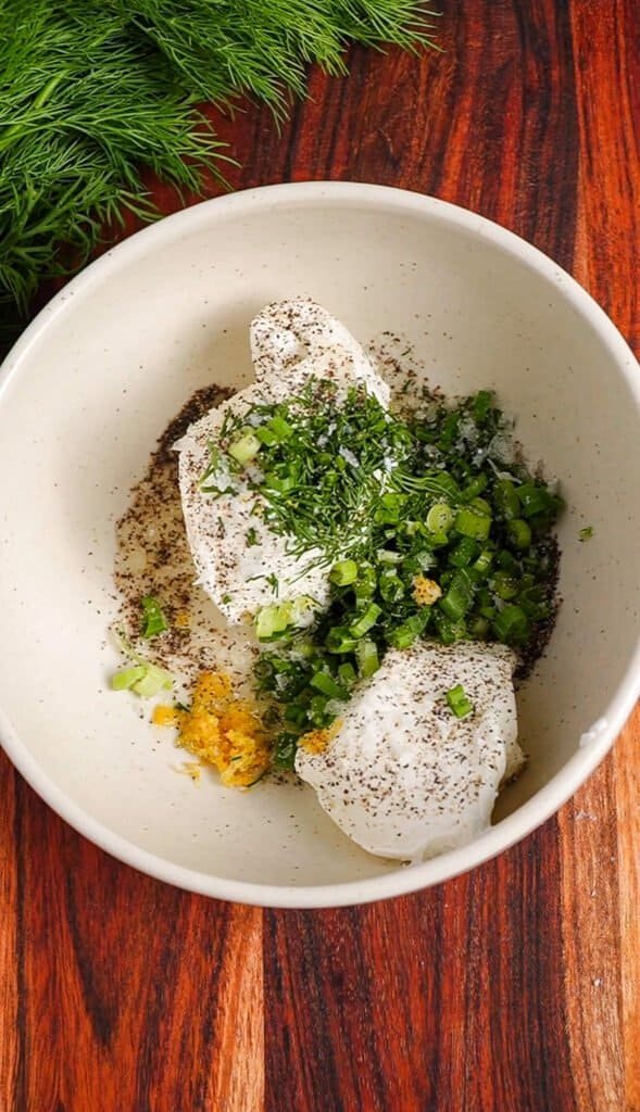 Labneh in a white bowl with chopped dill, chives, lemon zest, and black pepper before mixing