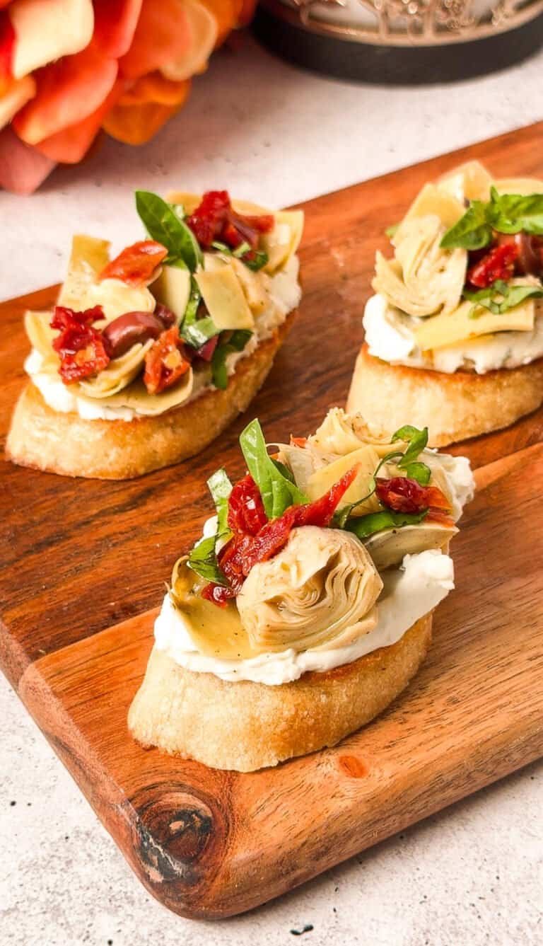 Three artichoke crostini with sun-dried tomatoes and olives arranged on a wooden serving board