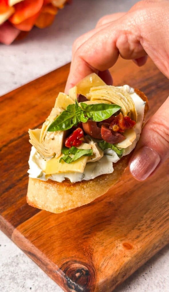 Hand holding an artichoke crostini topped with sun-dried tomatoes, olives, and basil on labneh