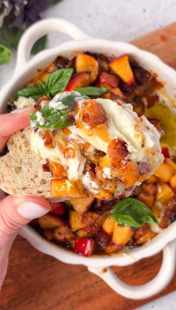 Crostini dipped into baked peach bacon dip with melted cheese and fresh basil