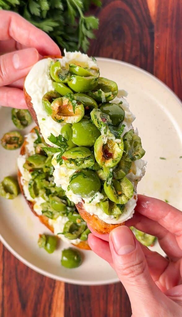 Hand holding a burrata toast piled with marinated Castelvetrano olives