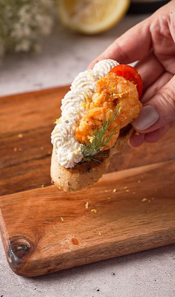 Whipped crab crostini with seared shrimp, cherry tomato, and lemon zest held above a wood board