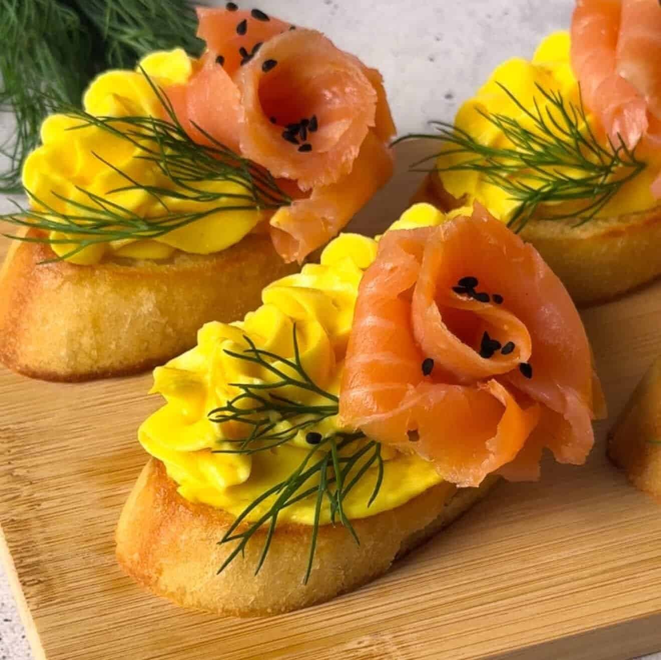 Finished smoked salmon crostini arranged on a wooden board, topped with golden egg–cheddar mousse, dill, and black sesame seeds.