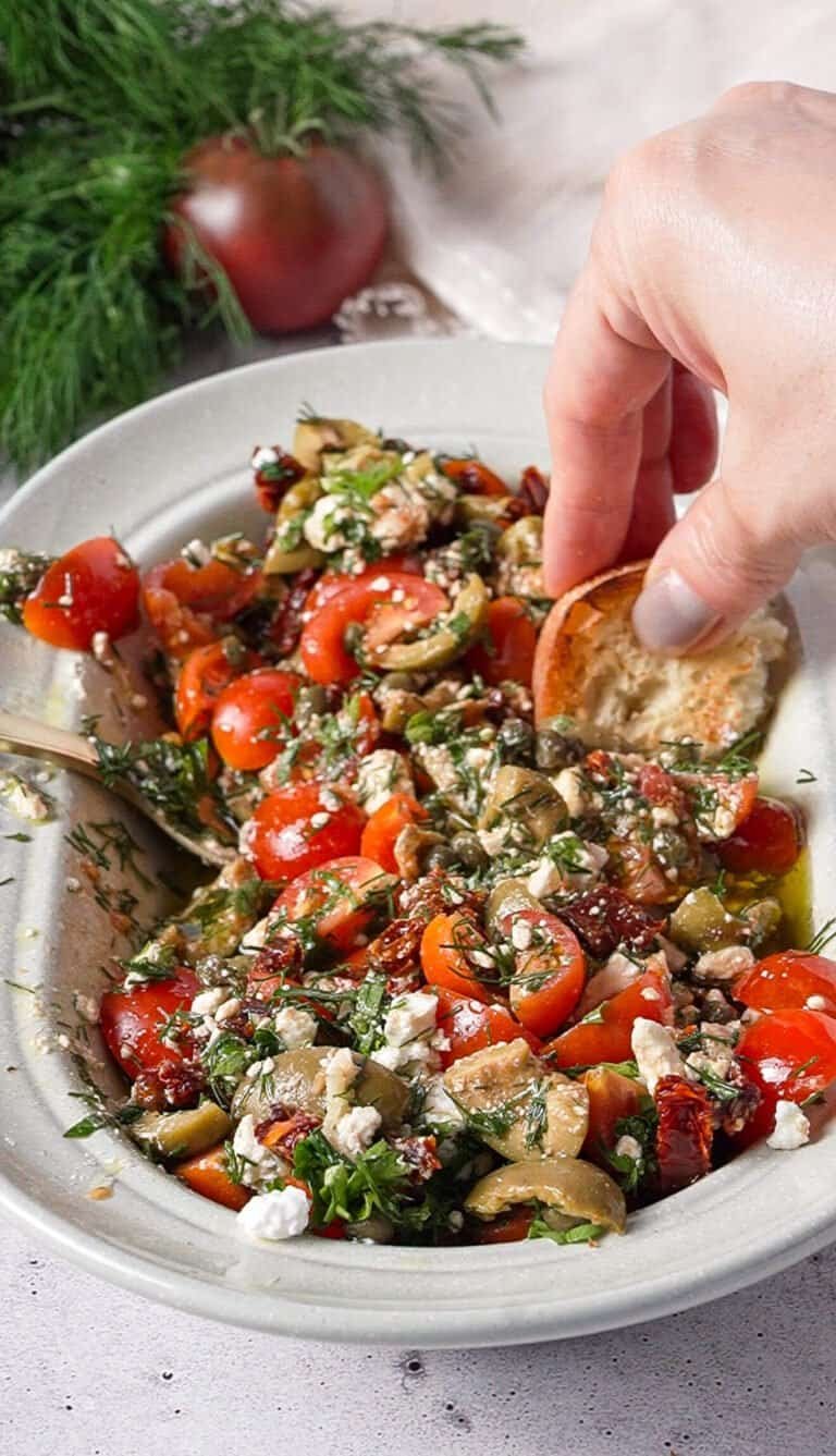 Hand dipping crusty bread into marinated feta dip with olives, tomatoes, and capers