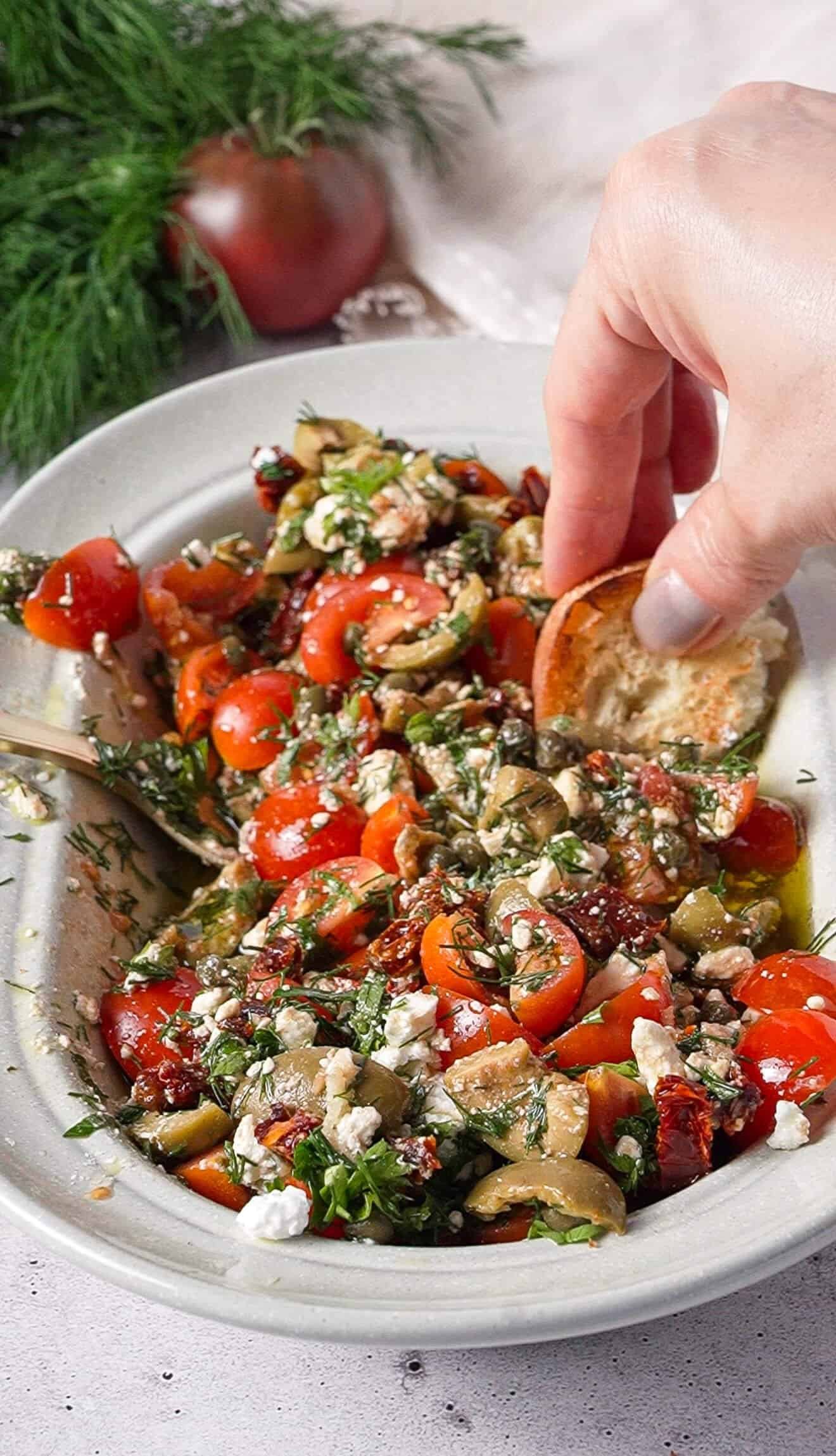 Hand dipping crusty bread into marinated feta dip with olives, tomatoes, and capers
