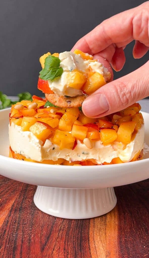 Hand lifting a cracker with peach and honey-mint goat cheese from the appetizer stack
